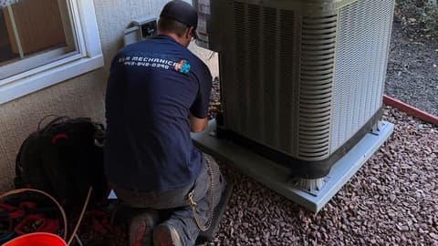 E&A technician in a navy company shirt kneeling at an outdoor condensing unit beside a house, tool bag and bucket on the gravel